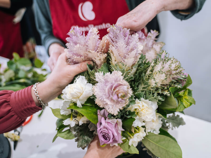 Close up of two pairs of hands working together to create a flower arrangement with purple and cream carnations